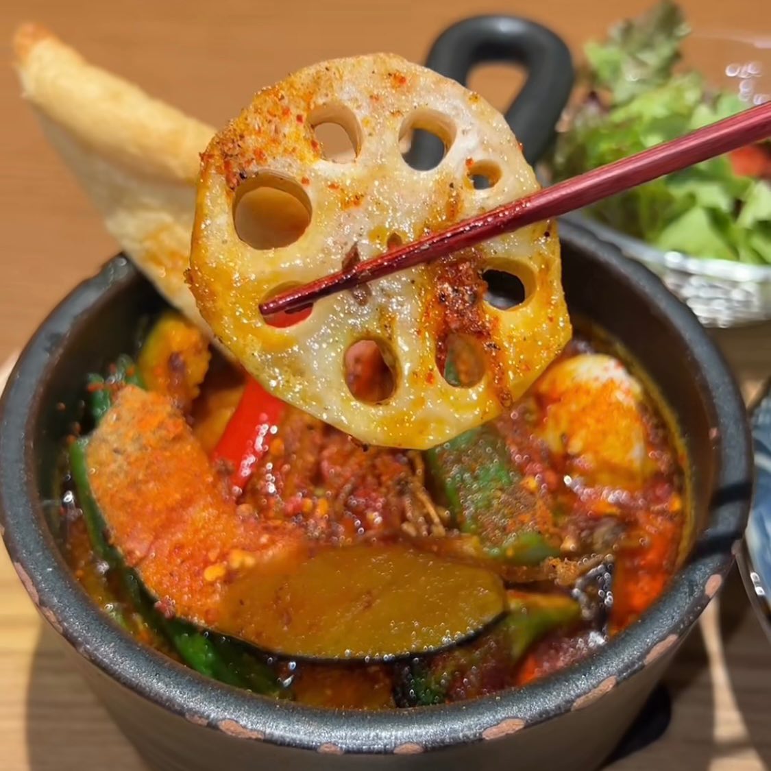 赤いスープのスープカレー。スパイスまみれのレンコンを箸で持ち上げ、茄子やかぼちゃなどの野菜が鍋に見えています。