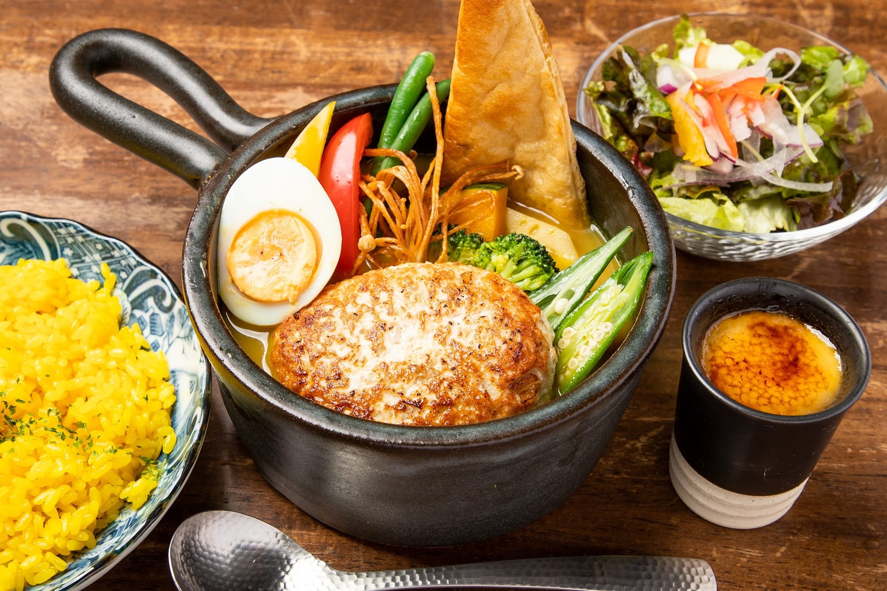 片手鍋に入ったハンバーグのスープカレー。ゆで卵、パンの角、オクラが入り、黄色いピラフとサラダが木のテーブルに並んでいます。