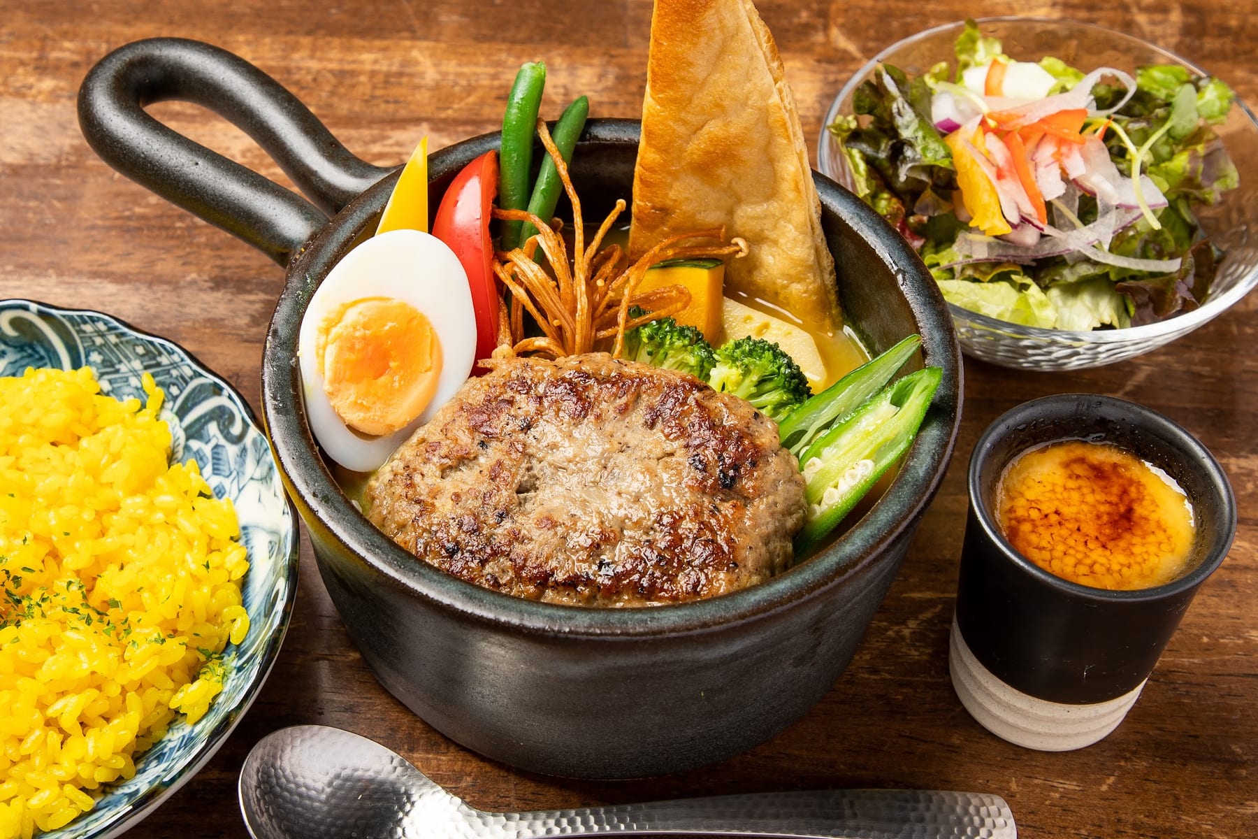片手鍋のスープカレーに大きなハンバーグが入った料理写真。ゆで卵、パンの角、サラダ、黄色いピラフ、プリンが添えられています。