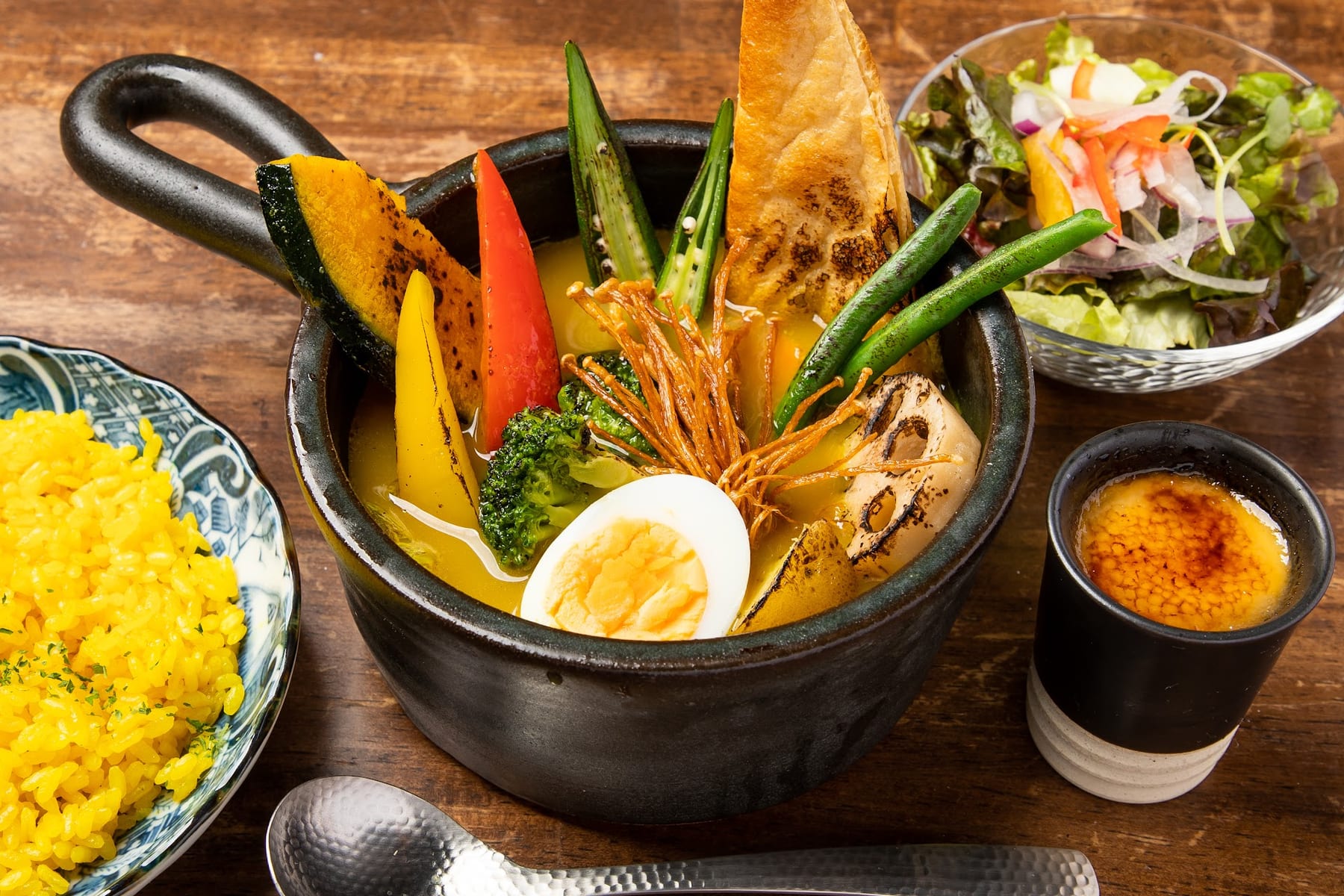鶏肉が入った黄色いスープカレー。ゆで卵、かぼちゃ、オクラ、パンの角が浮かび、ピラフ、サラダ、プリンが並んでいます。