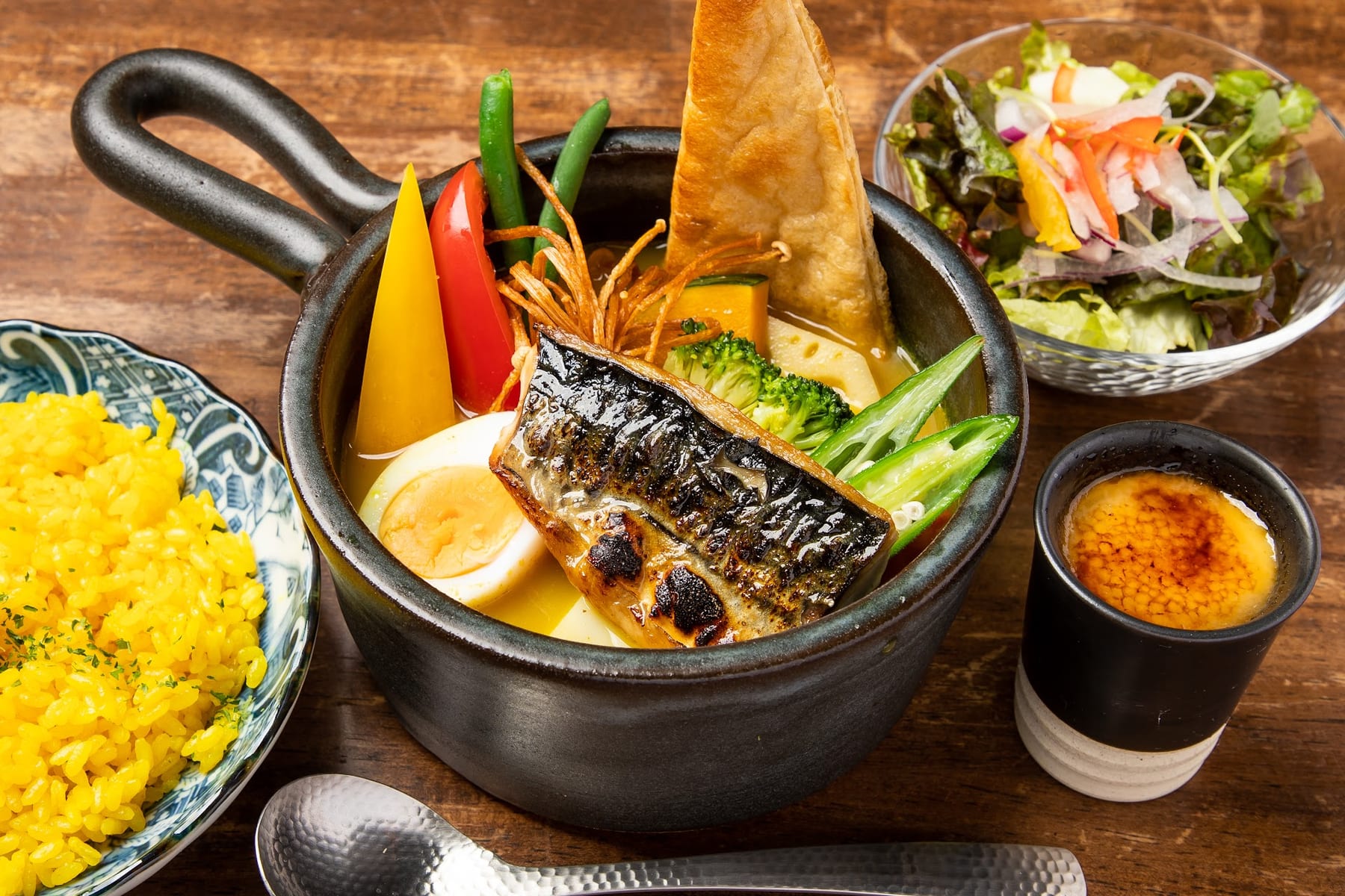 焼きサバがのったスープカレー。ゆで卵、パンの角、オクラが入った片手鍋に、黄色いピラフとサラダを添えた木のテーブルの構図です。