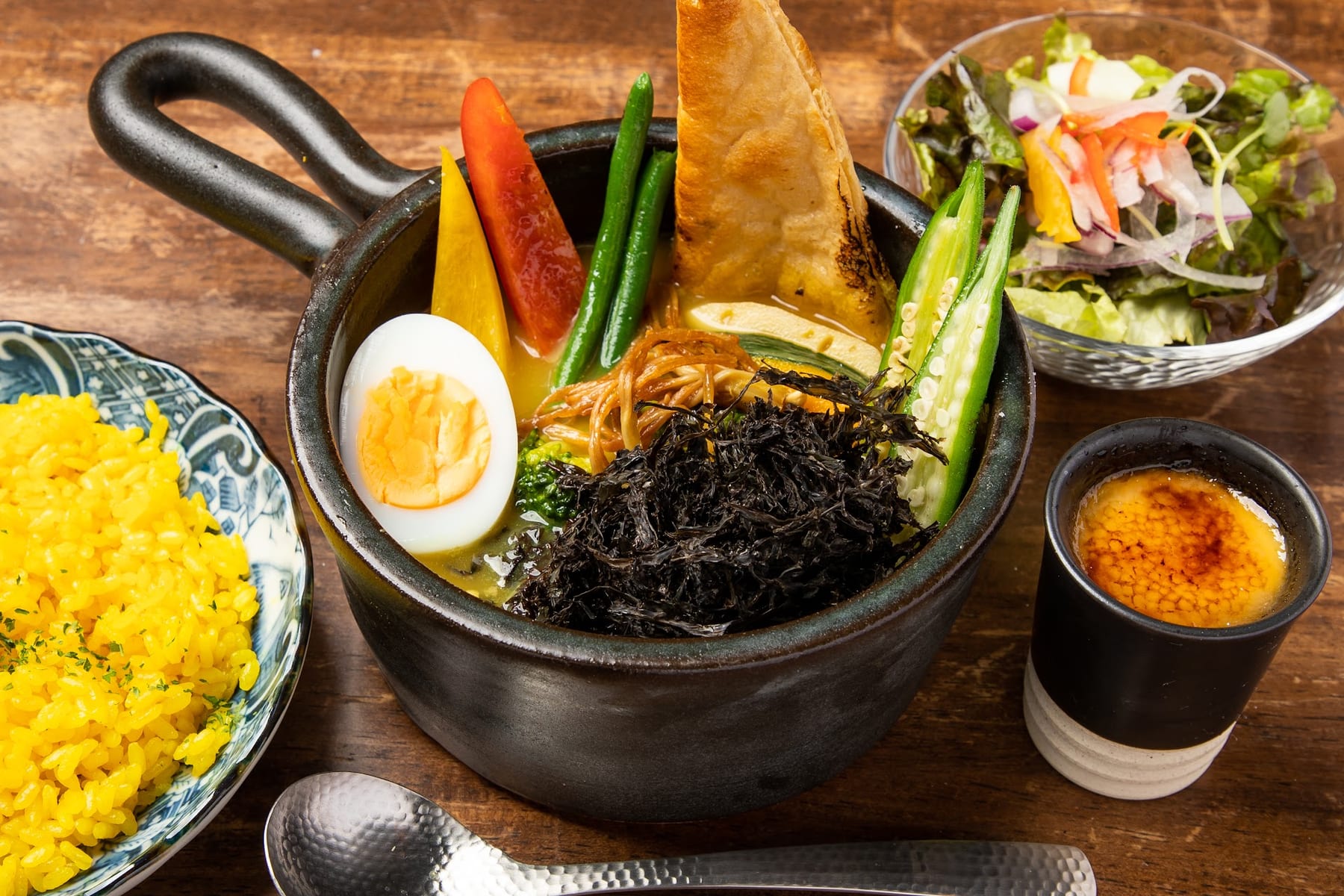 海苔や野菜がたっぷりのスープカレーを片手鍋で写した料理写真。ゆで卵、パンの角、オクラが浮かび、黄色いピラフやサラダとともに明るく配される。