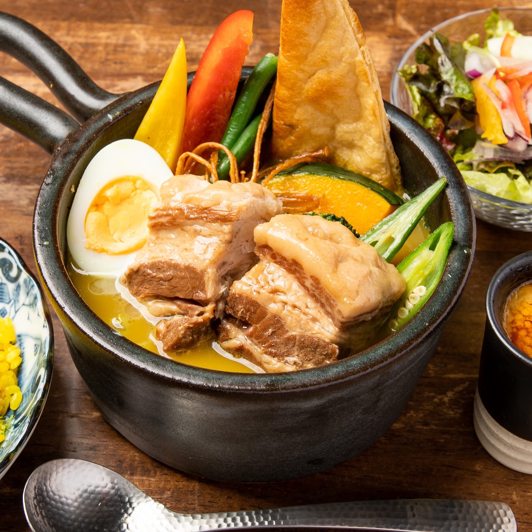 柔らかそうな豚の角煮が大きくのったスープカレーのクローズアップ。ゆで卵、かぼちゃ、オクラ、パンの角が入り、サラダとスプーンを添えた温かい画面。